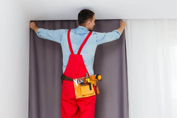Image of a man in a red uniform and equipment working on placing the gray curtain on the wall.