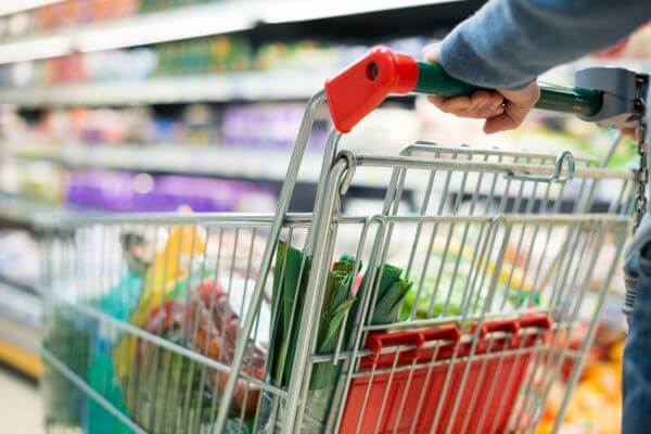 A picture of a cart with full of groceries stuff,  handled by a man's hand in a shopping store.