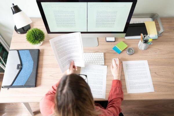 Image of the girl holding a paper with text and holding into the mouse while referring to the document in the personal computer.