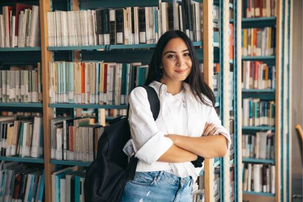 Picture of the  college student wearing a white sleeves and a bag standing along the book shelve.