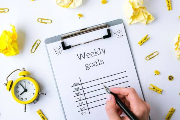 The picture of a weekly goals planner with a pen in hand and stuff in the surroundings.
