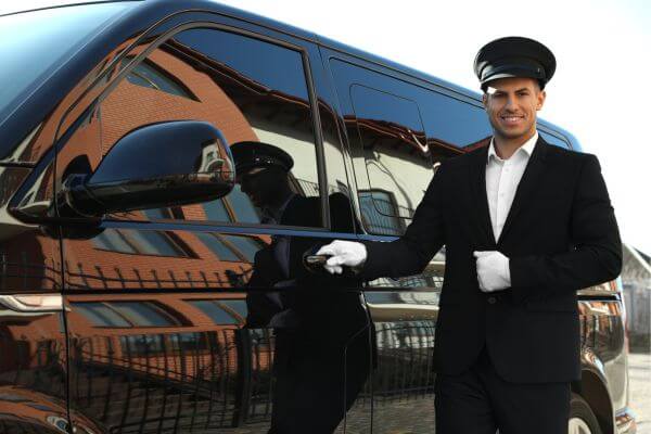 Image of the valet driver in a black and white suit, cap, and white gloves in a black car behind.