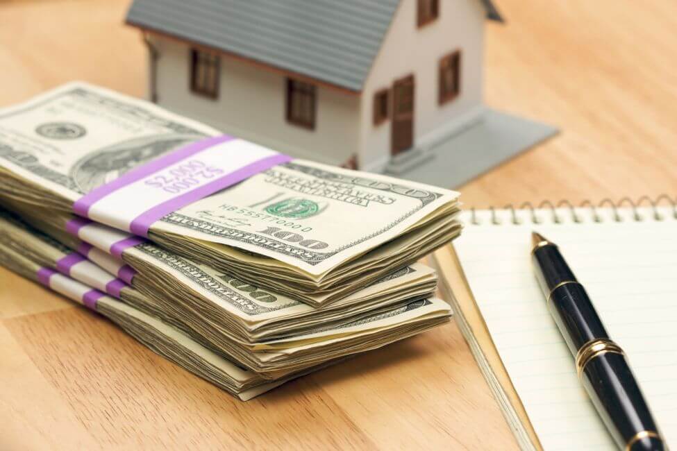 Picture of the dollar bill, house, pen, and notebook on a wooden desk for how much should I save before moving out.