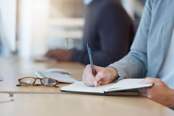 Picture of the man writing and holding a pen in a notebook, eyeglasses, and keyboard on the desk.