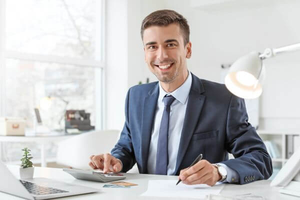 Image of a man in a suit holding a pen and tapping the calculator button and a laptop in a desk.