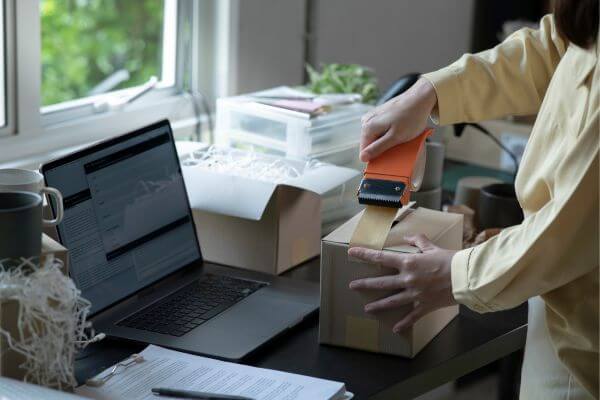 Picture of the woman packing a stuff in a box and taping to it secure, also a laptop in a desk and surrounded by a stuffs.