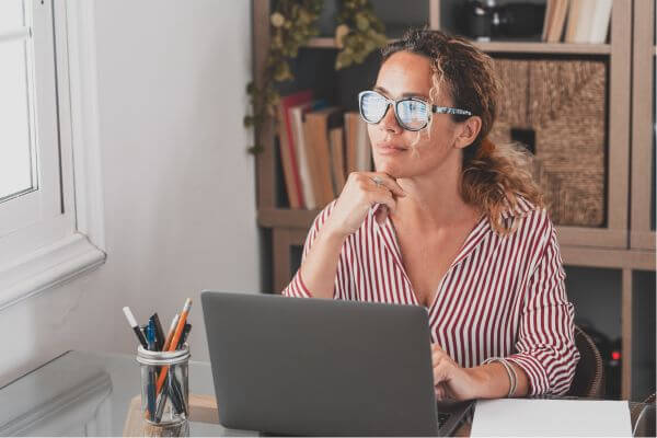 Picture of the woman thinking deep while holding into her gray laptop and a pens in her desk.