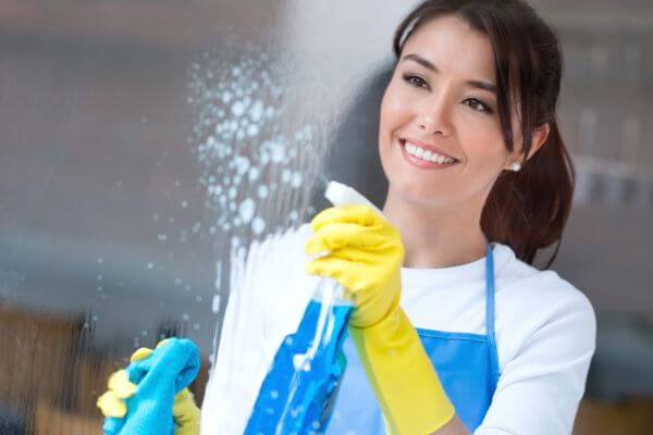 Image of a woman wearing apron and a yellow gloves while cleaning the mirrored window. 