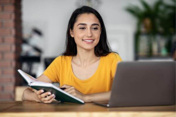 Picture of woman holding a pen and  a notebook while looking at the laptop.