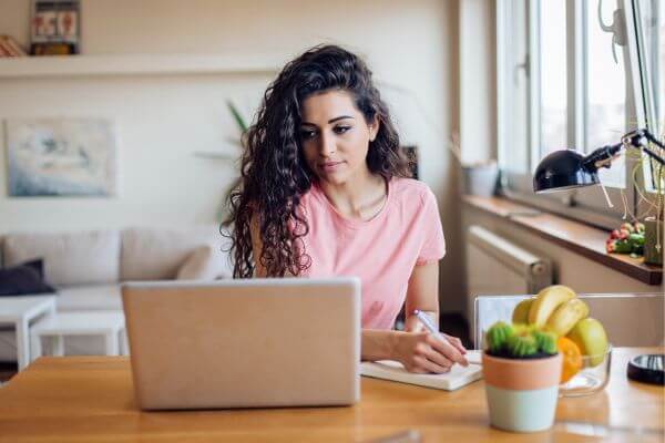 Picture of  woman writing something in a notebook while looking at the laptop.