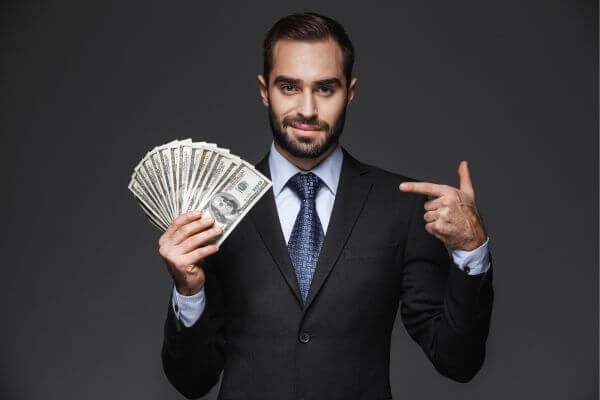Image of a man in a suit holding  a dollar bill while pointing to it. 