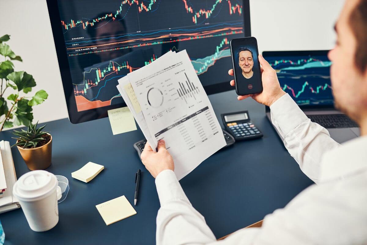 The picture of the man using a personal computer and holding a paper and phone having a conversation with the man online indicating to trade stocks and grow income over time.