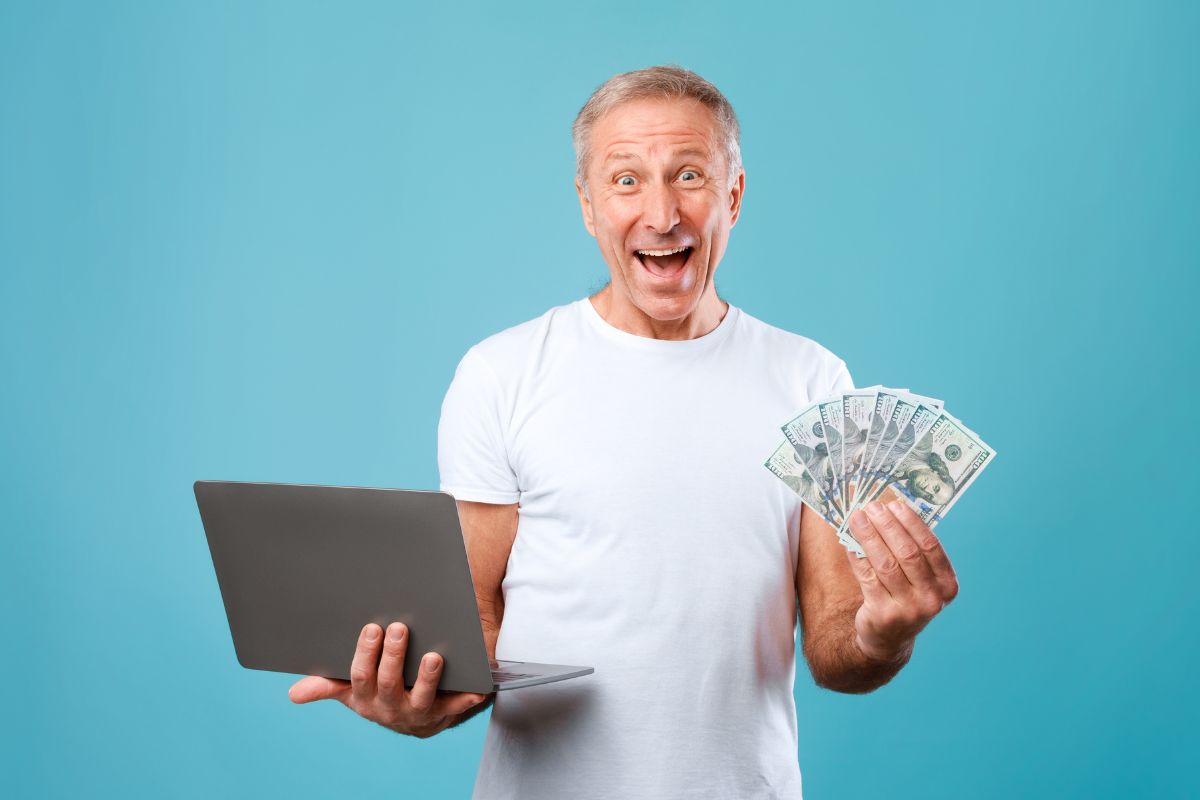 An aging man holding a laptop and money.
