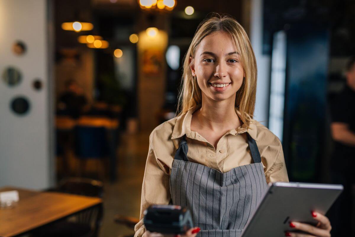 The image of the teen holding a tablet device standing in the cashier area means working a part-time job in food service to gain experience and earn regular income.