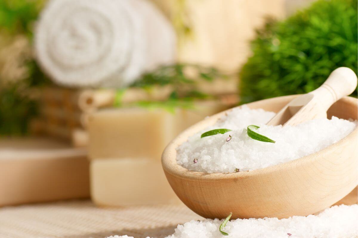 The picture of a towel, soaps, and a wooden bowl with salt, leaves and scoop referring to make a DIY bath salts as a gift that is thoughtful and perfect for pampering friends or family without a high price tag.
