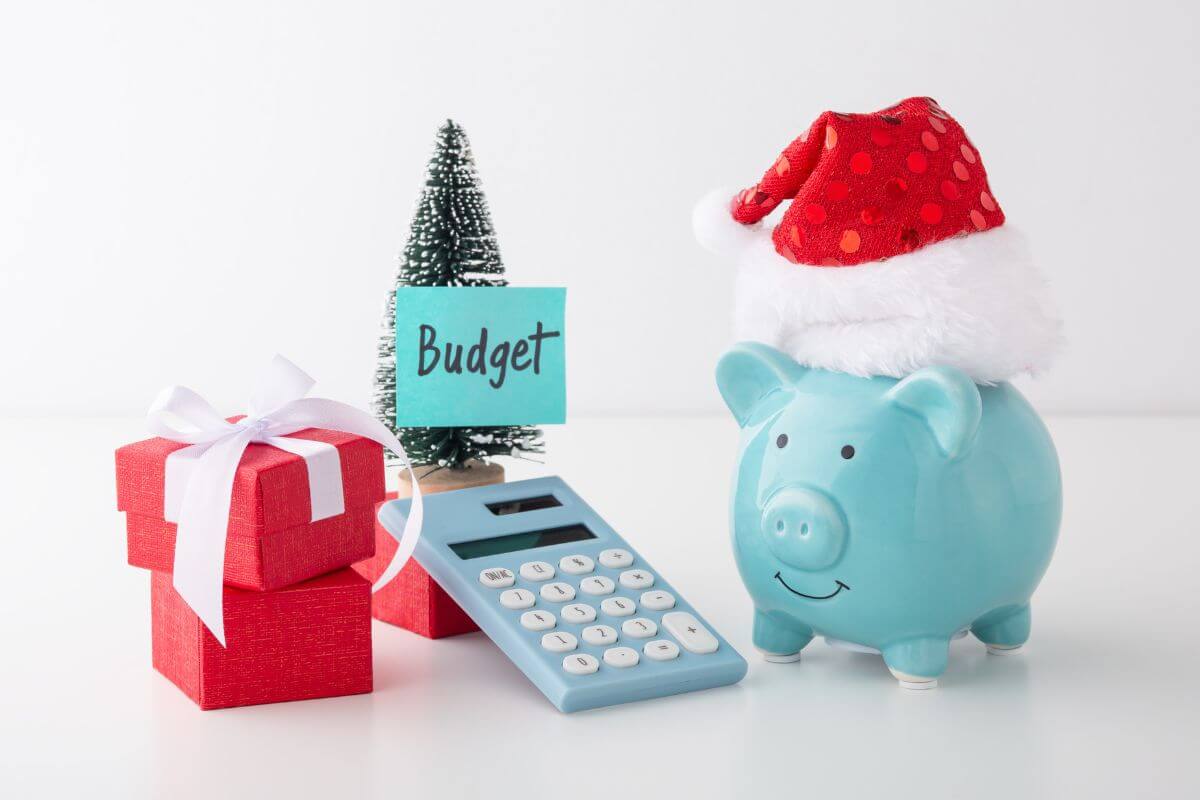 The photo of mini gifts, a mini tree with a note on the paper, a calculator, and a piggy bank with a hat indicate to set a strict holiday budget and stick to it which helps you manage expenses and avoid debt.