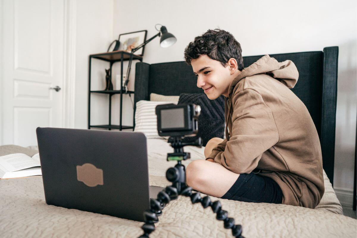 The picture shows a teen filming himself while using a laptop indicating to start a blog on a topic you enjoy that might turn into a profitable hobby.