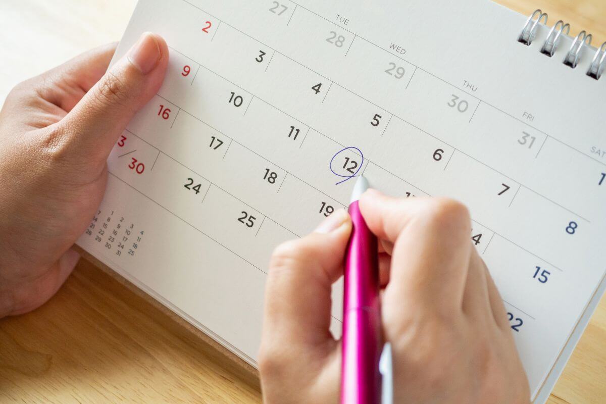 The image shows a girl's hand holding a calendar and a pen writing on it which refers to utilizing a shared calendar to coordinate family and work commitments that help prevent scheduling conflicts.