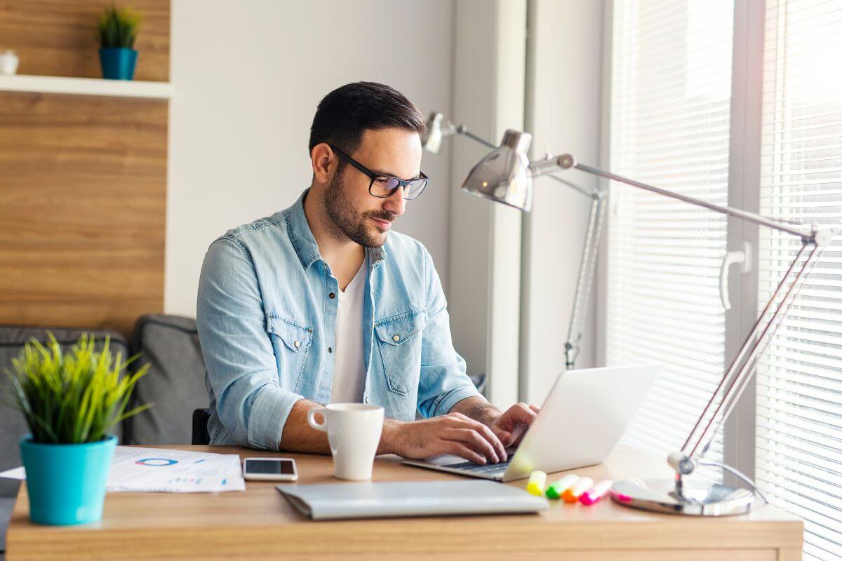 The image shows a plant, lamp, papers, cup, and a man using a laptop indicating to consider flexible work like remote work that offer more control over your schedule.