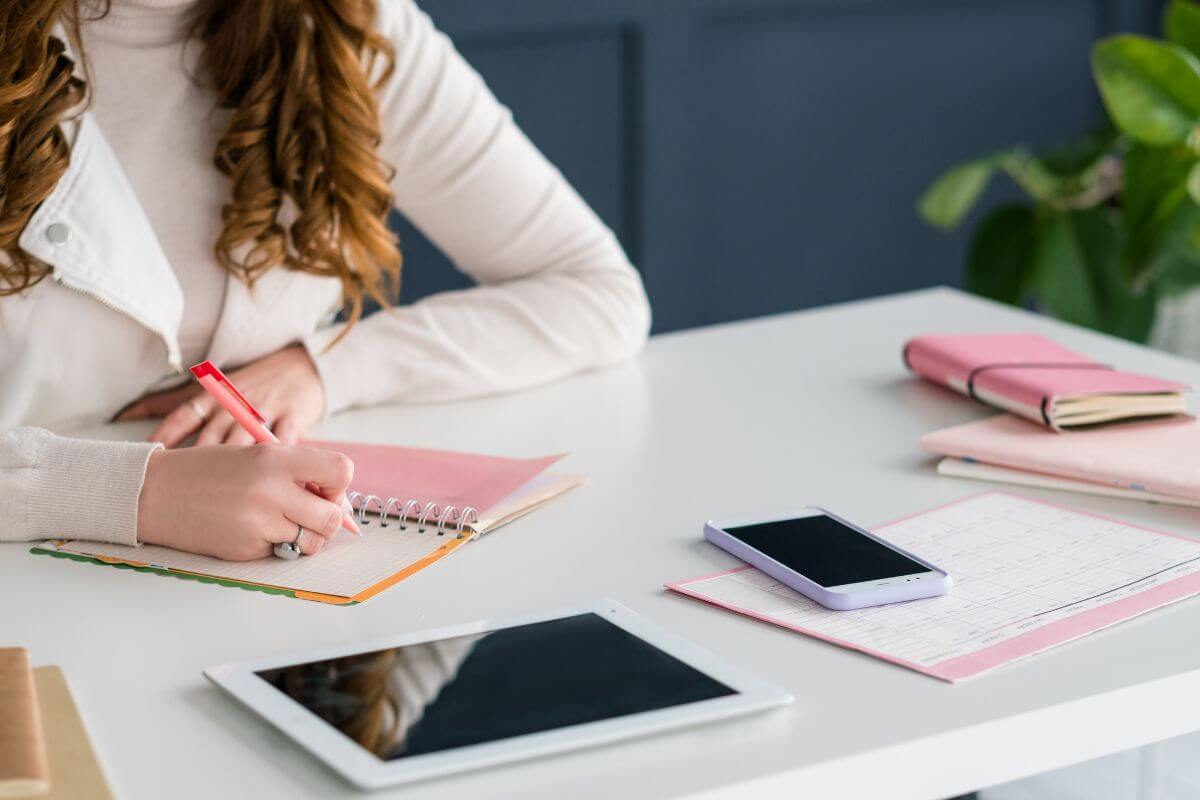 The image shows a tablet, a phone, papers, notebooks, and a girl's hand and writing on the notebook indicating to manage social media accounts on small businesses that lets you work remotely and build your marketing skills.