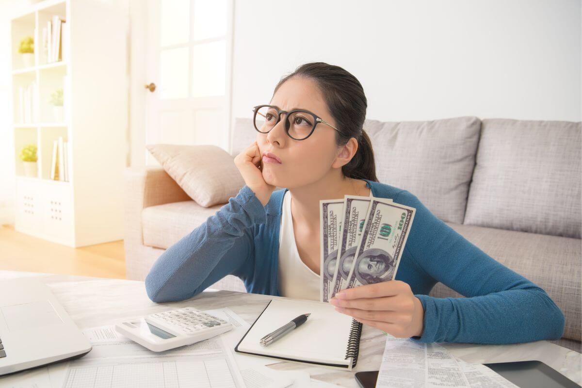 The picture tells about a laptop, a calculator, a notebook with a pen, and a girl thinking while holding dollar bills which means cutting back on discretionary expenses and regaining control of our finances.