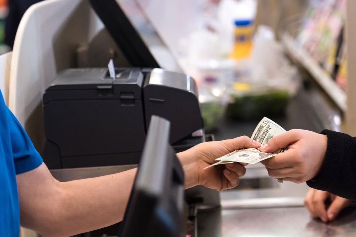 The image shows a customer paying the cashier with cash indicating to use cash for purchases to avoid more debt and stay within your means. 
