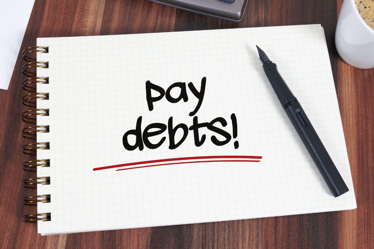 The image shows a coffee, a pen, and a notebook with notes referring to prioritizing paying off high-interest debt first can frees up more cash for other goals.