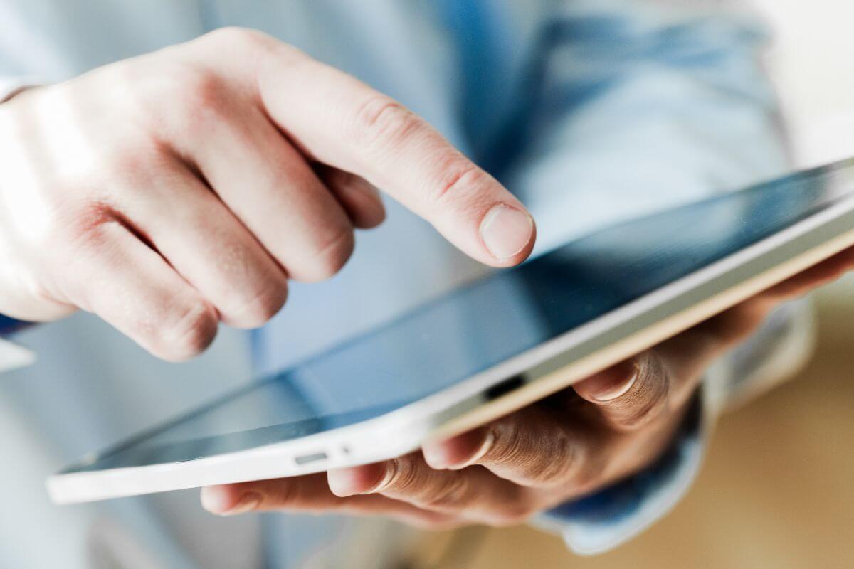 The image shows a man's hand using a tablet device which means reviewing and adjusting your monthly subscriptions to reduce unnecessary expenses.