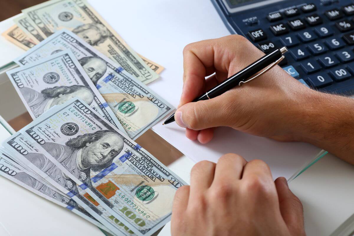 The photo shows a calculator, dollar bills, and a man's hand and writing on the paper which refers to setting financial goals for the next few months that help you stay motivated and focused on recovering after the holidays.