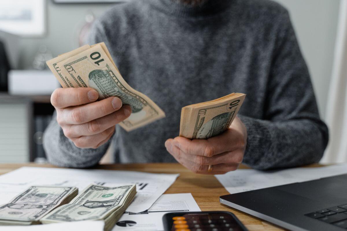 The image shows a laptop, papers, a phone with a calculator, and a man dividing the dollar bills into groups which refers to reevaluating your financial priorities and adjusting as needed to better align with your goals.