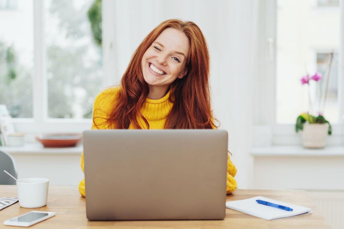 The picture shows a phone, a cup, a notebook with pen, and a girl using a laptop to search while winking indicating to get on track to be a millionaire with no money and financial independence.