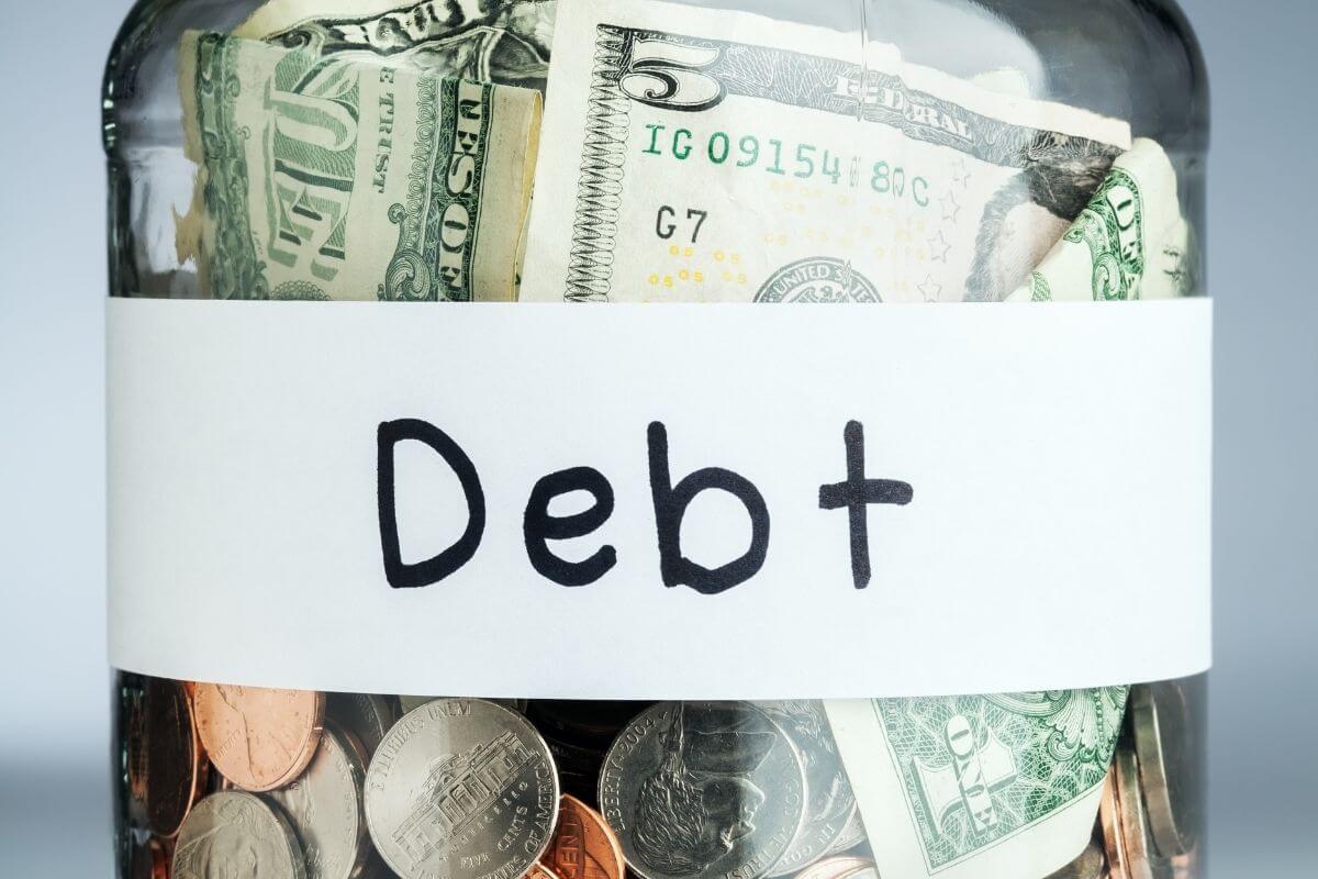 The image tells about a jar filled with dollar bills and coins with a white note referring to unsecured debt exceeding your savings as a warning sign you have too much debt.