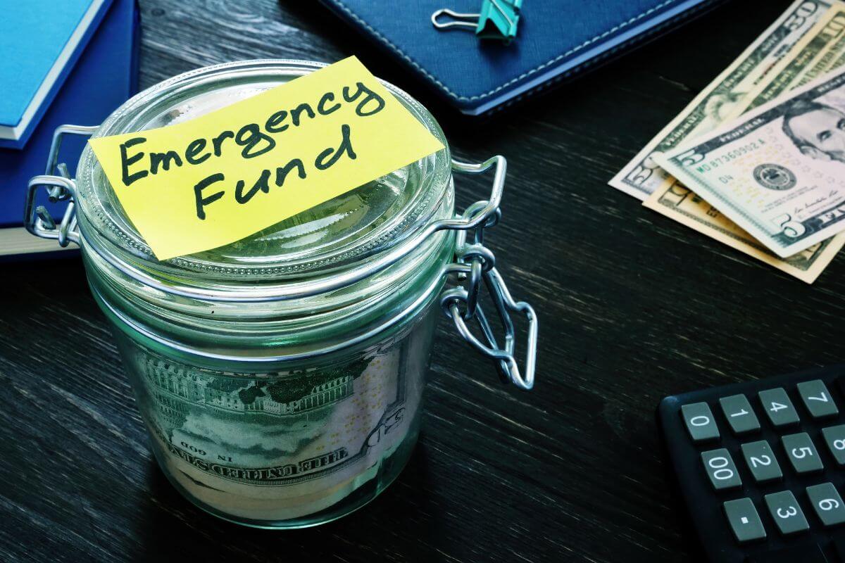 The picture shows a notebook, dollar bills on the table, a calculator, a jar with dollar bills inside, and a piece of paper with a note referring to debt covering emergency is a red flag that drowns you on unexpected expenses.