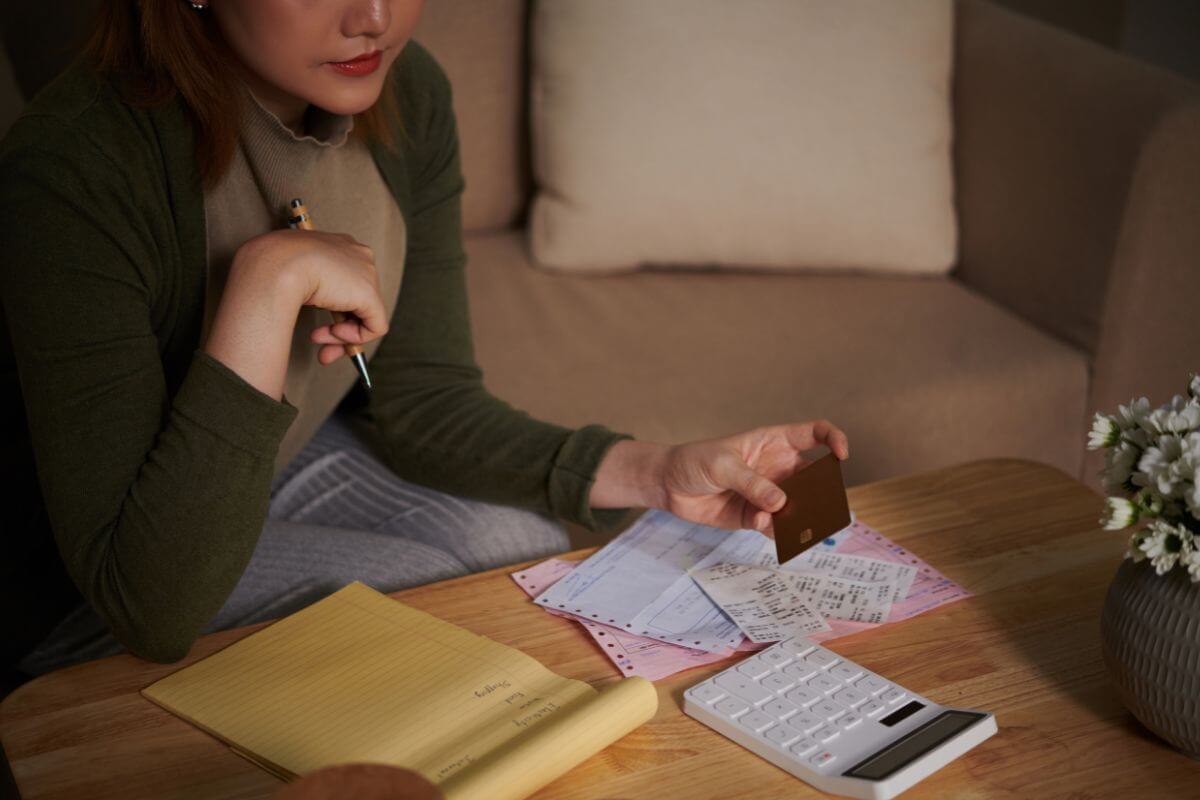 The photo of papers on the table, a calculator, and a girl holding a card and a pen which means too much debt can lead to struggling to meet loan and credit card payments, adjust your budget, and seek help if needed.