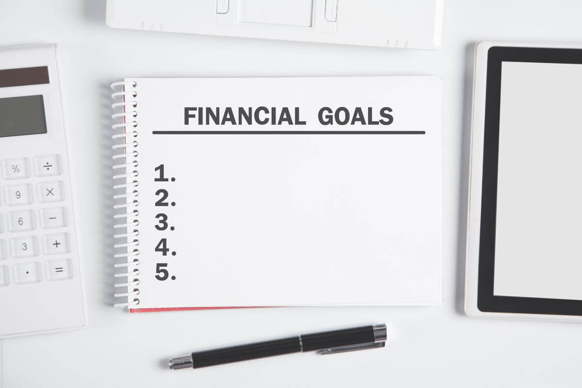 The picture tells about a calculator, a pen, and a notebook with notes which refers to setting small and achievable financial goals that help you feel more confident and make money management feel doable.