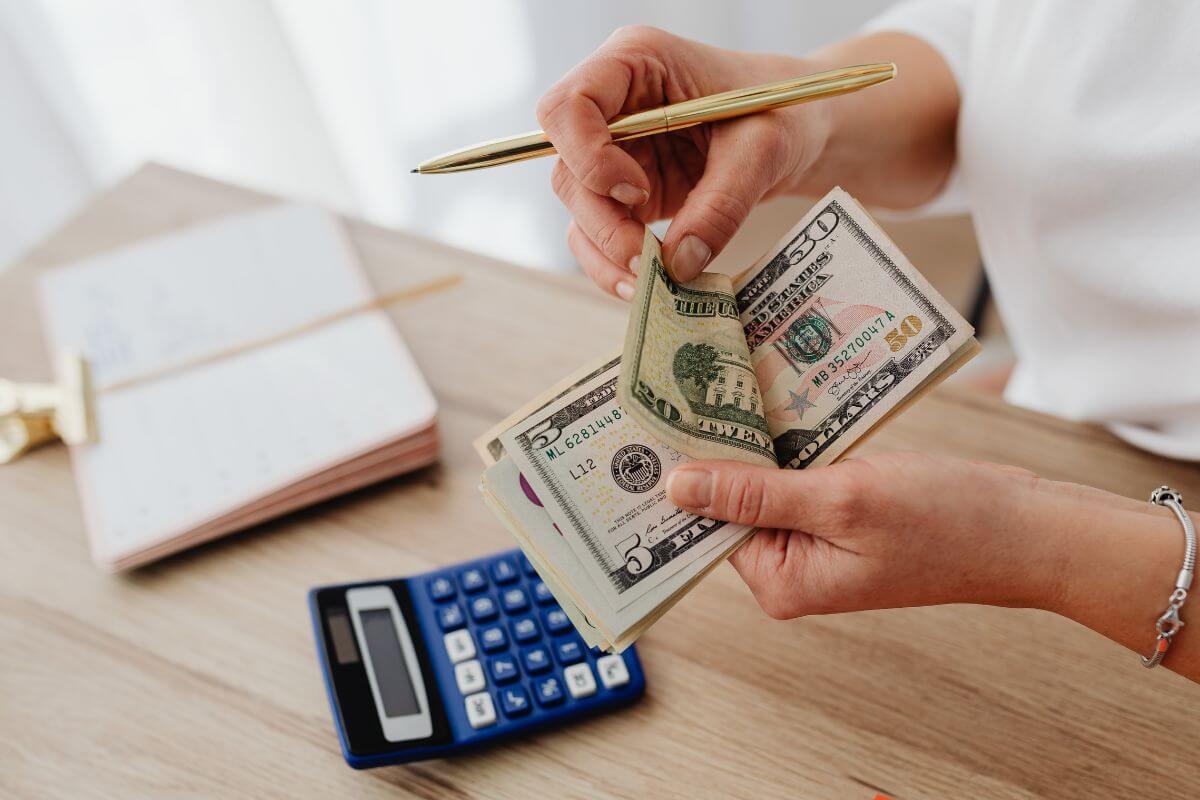 The picture tells about a notebook, a calculator, and a girl's hand counting money with a pen which means understanding that everyone's financial journey is different and knowing that others face similar struggles can help you feel less isolated.