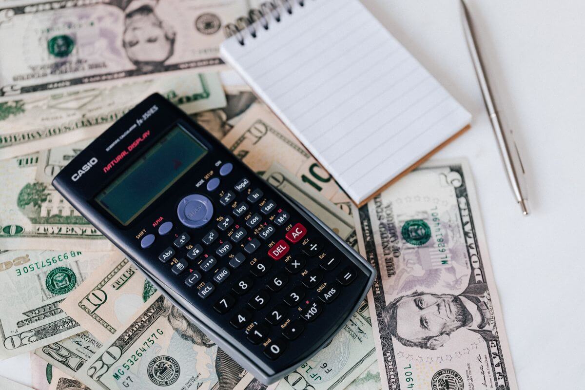 The photo shows a pen, a calculator, dollar bills, and a mini notebook which means creating a detailed budget to track income and expenses that helps you focus on paying off debt faster.