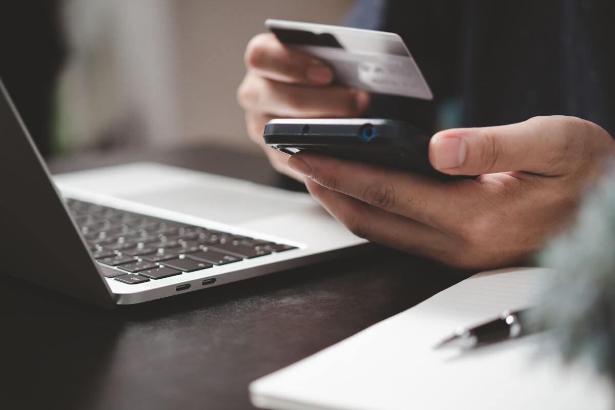 The image shows a laptop, a pen, a notebook, and a man's hand holding a phone and a credit card indicating to automate debt payments to ensure consistency that keeps you on track and avoids missed deadlines.