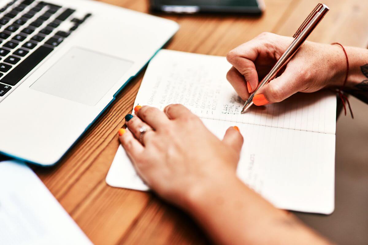The picture shows a laptop and a girl's hand and writing on the notebook indicating to stay motivated by tracking progress and regularly checking how much debt you've paid off.