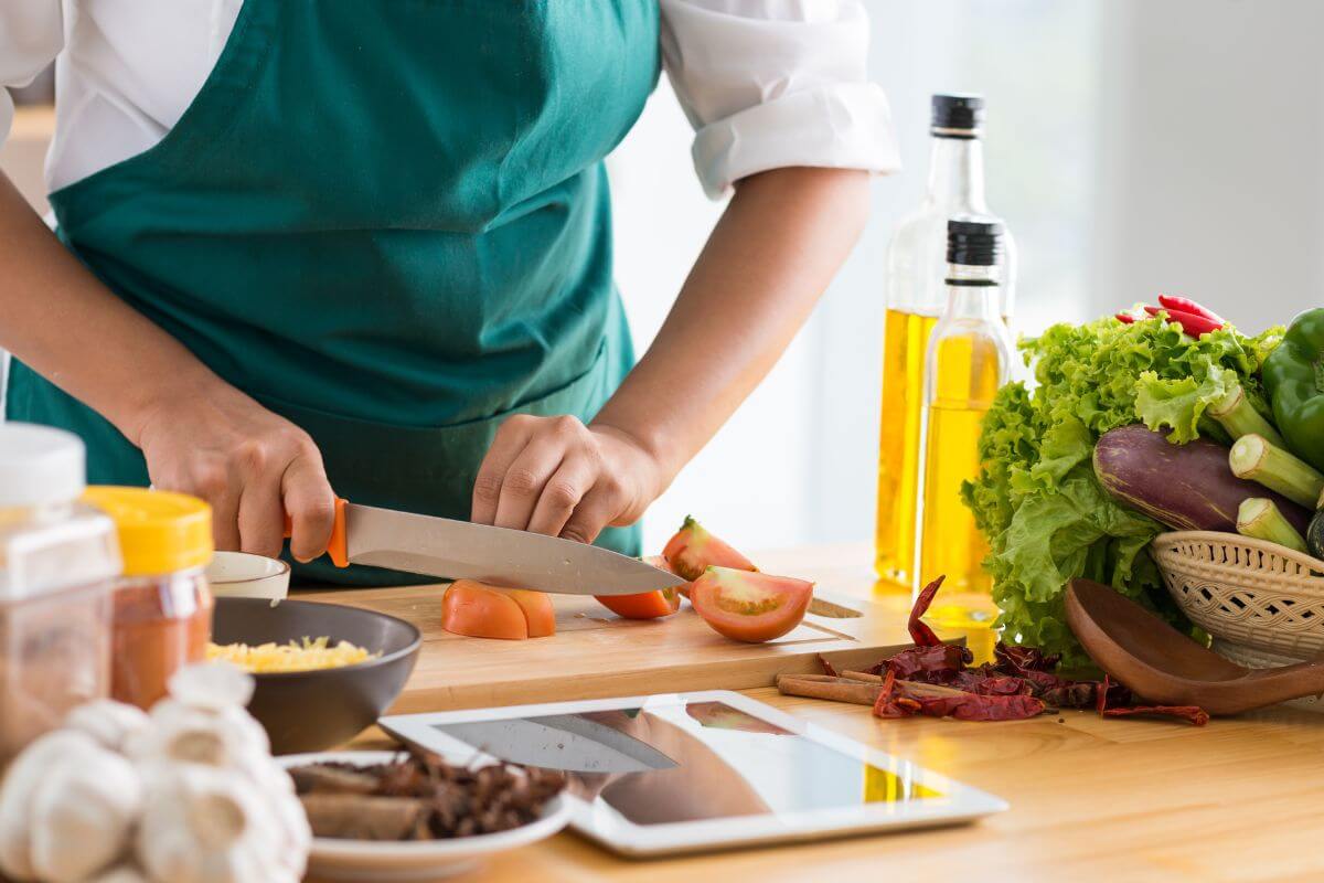 The photo of ingredients, oil, seasonings, a tablet device, and a girl's hand chopping tomatoes indicates to cook meals at home and avoid dining out to save money fast and stretch your grocery bill even further.