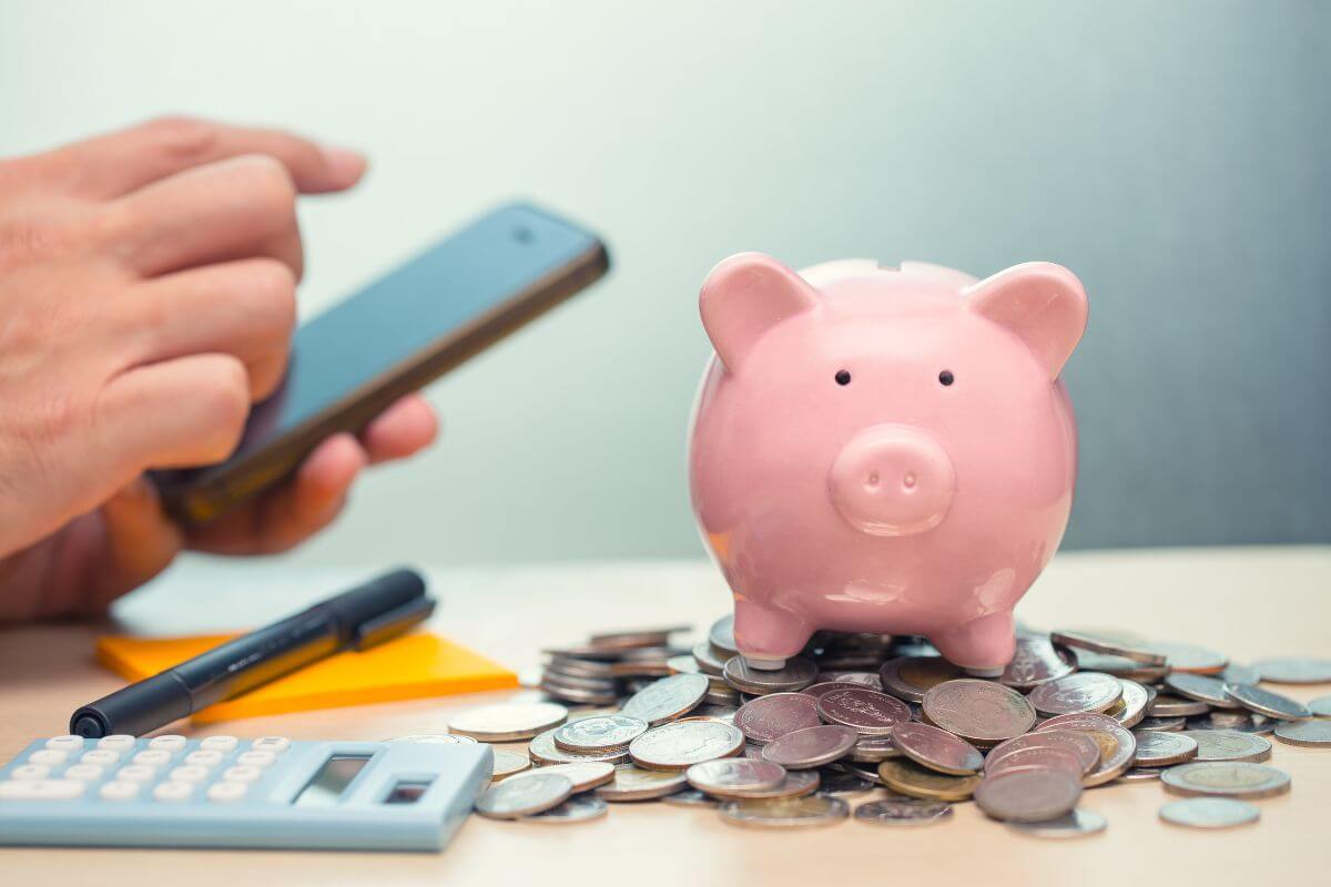 The photo tells about coins, a piggy bank, a calculator, a pen, sticker notes, and a man's hand using a phone indicating to use a budgeting app to stay on track and set limits that help you avoid overspending and hit your financial goals.