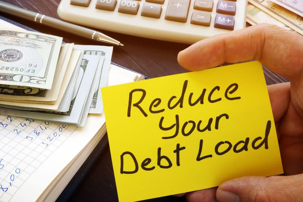 The picture shows dollar bills, a calculator, a pen, a notebook, and a man's hand holding a yellow paper with notes referring to reducing or eliminating debt frees up money for saving or investing and stability in your life.
