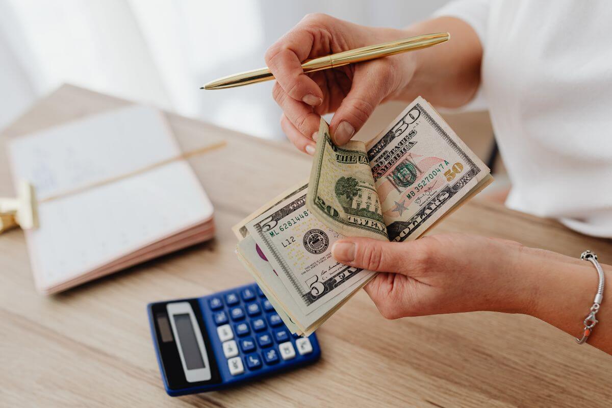 The photo tells about a notebook, a calculator, and a girl's hand counting money with a pen in her hand indicating that automating extra payments to debt accounts helps you stay on track without overthinking it.