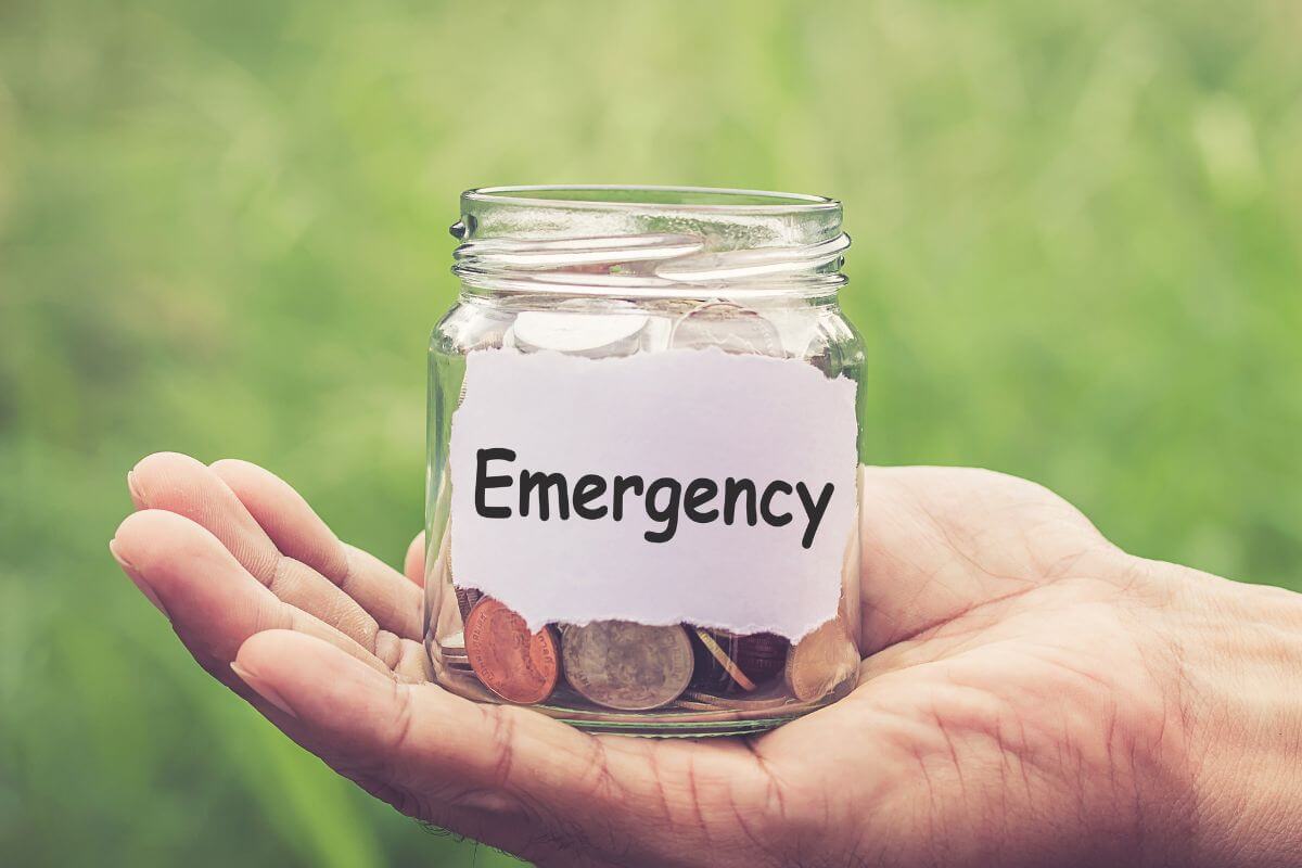 The photo of a man's hand holding a jar filled with coins and with a note referring to starting an emergency fund, even if small as a cushion for unexpected costs.