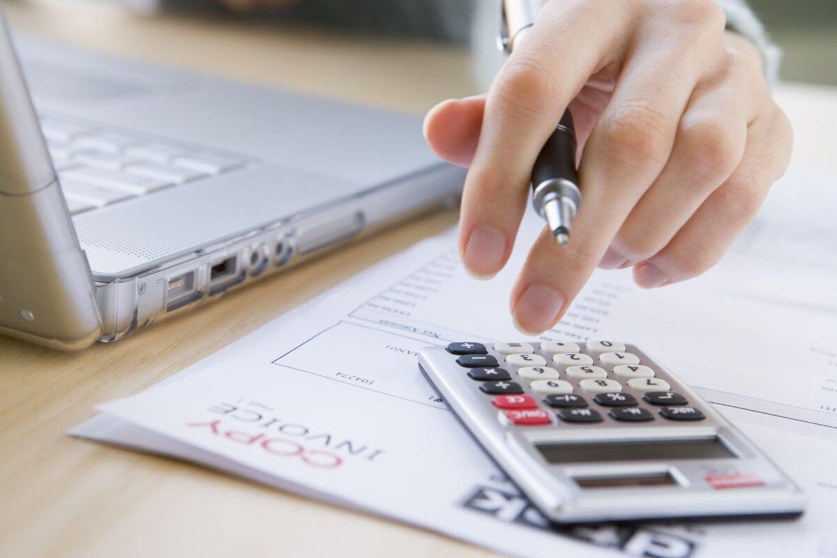 The image shows papers on invoices, a laptop, and a girl's hand using a calculator that automates bill payments and savings so you won't worry about missed payments or forgetting to save.