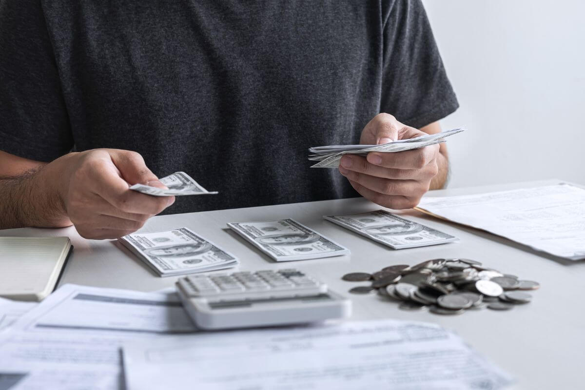 The photo shows papers on the table, coins, a calculator, a notebook, dollar bills divided into groups, and a man's hand holding dollar bills indicating to track your income and expenses weekly to stay in control without getting overwhelmed by too much data.
