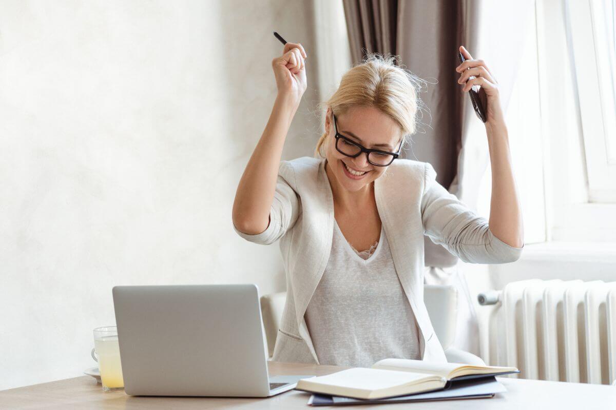 The image of a laptop, a glass of juice, a notebook, and a girl happy and cheering which refers to celebrating small financial wins keeps you motivated and reminds you that progress is happening.