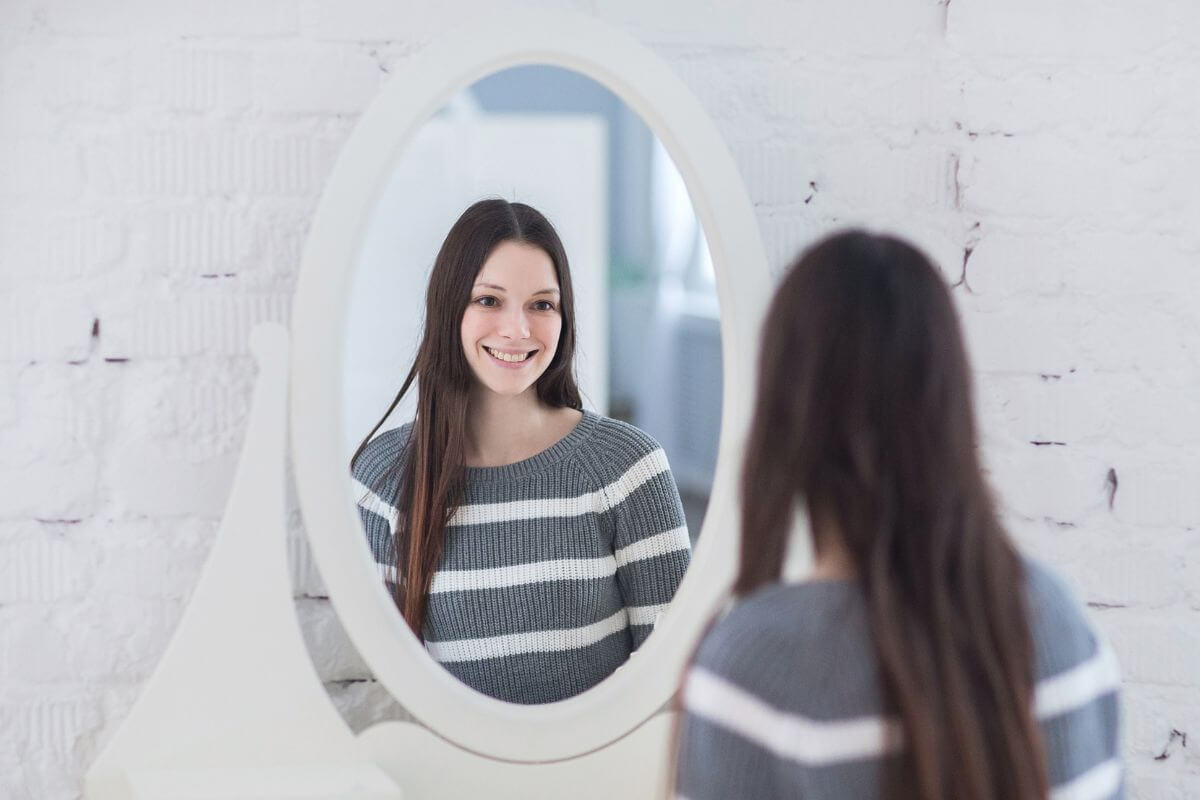 The image shows a girl looking at the mirror and smiling which means to remind yourself that improvement takes time and trust that you're moving in the right direction.