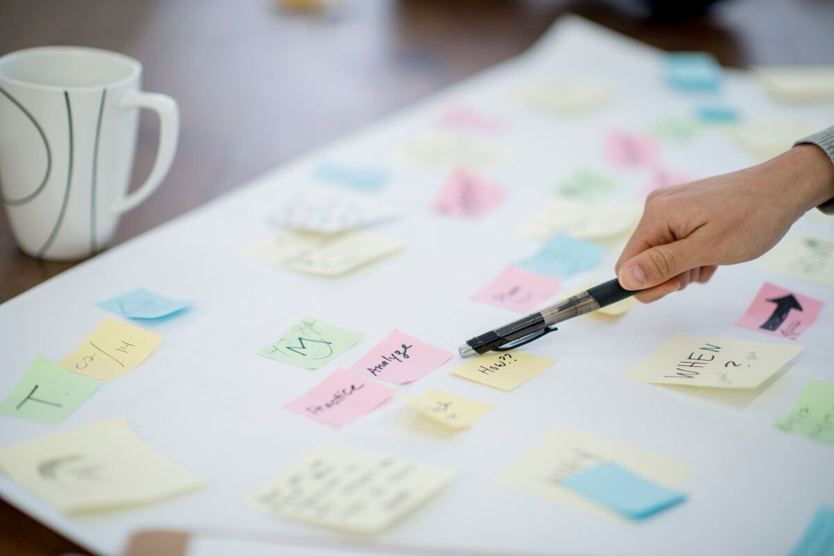 The photo of a cup and a board with sticker notes pointed by a girl's hand with a pen indicates that creating a vision board for financial goals keeps you motivated and focused on the future.