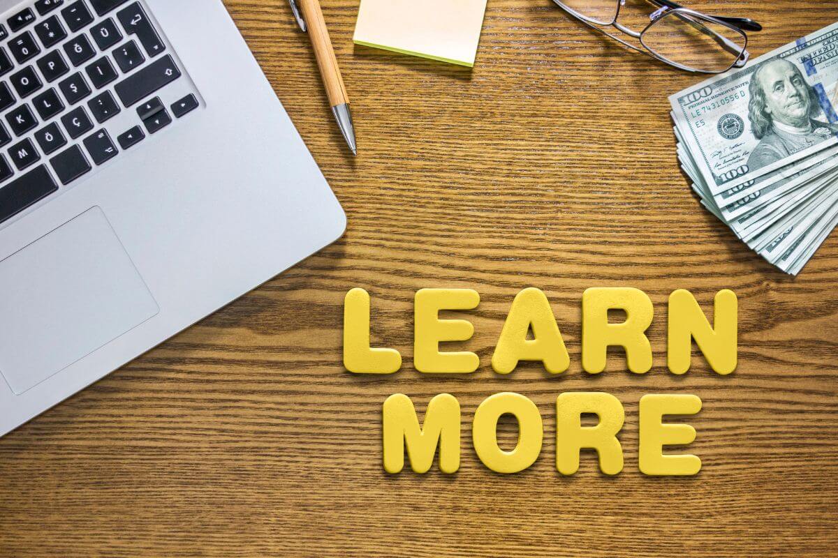 The image shows a laptop, an eyeglass, dollar bills, a pen, sticker notes, and letters of learn more which means to find more ideas to motivate yourself to be better with money to keep you on track and reach your financial goals.